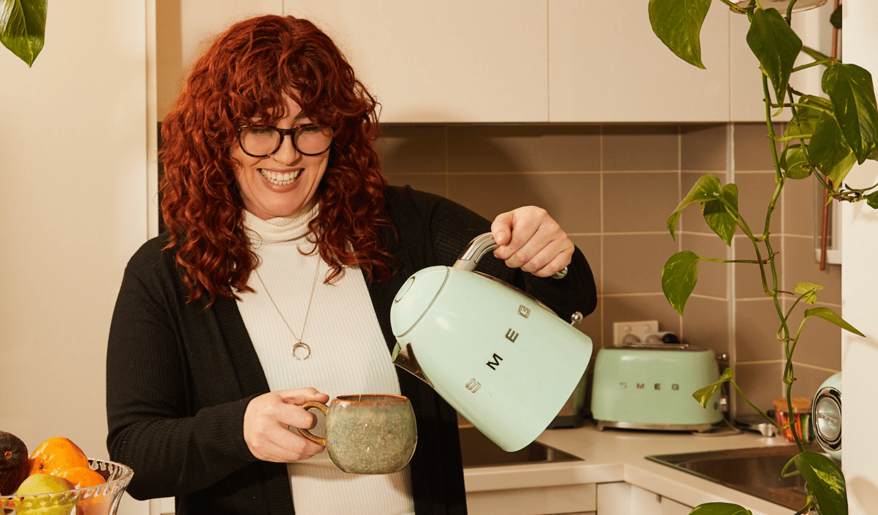 A smiling woman is pouring tea from her green teapot into her green mug. In the background, there's a plant, a sink, a green toaster, and a fruit plate. The walls are painted white.