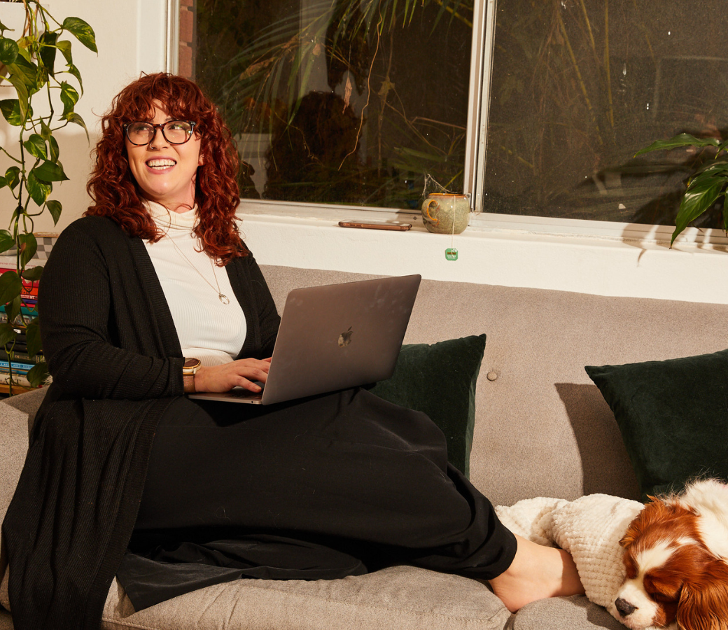 A person typing on a laptop sits on a grey couch, smiling, while a dog sleeps next to them. A plant and books are in the background, near a window and a mug.