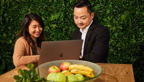Two people working together on a laptop at a table with a bowl of fruit, set against a green leafy background