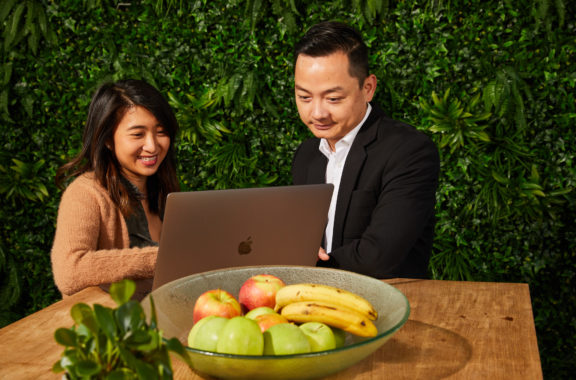 Two people working together on a laptop at a table with a bowl of fruit, set against a green leafy background