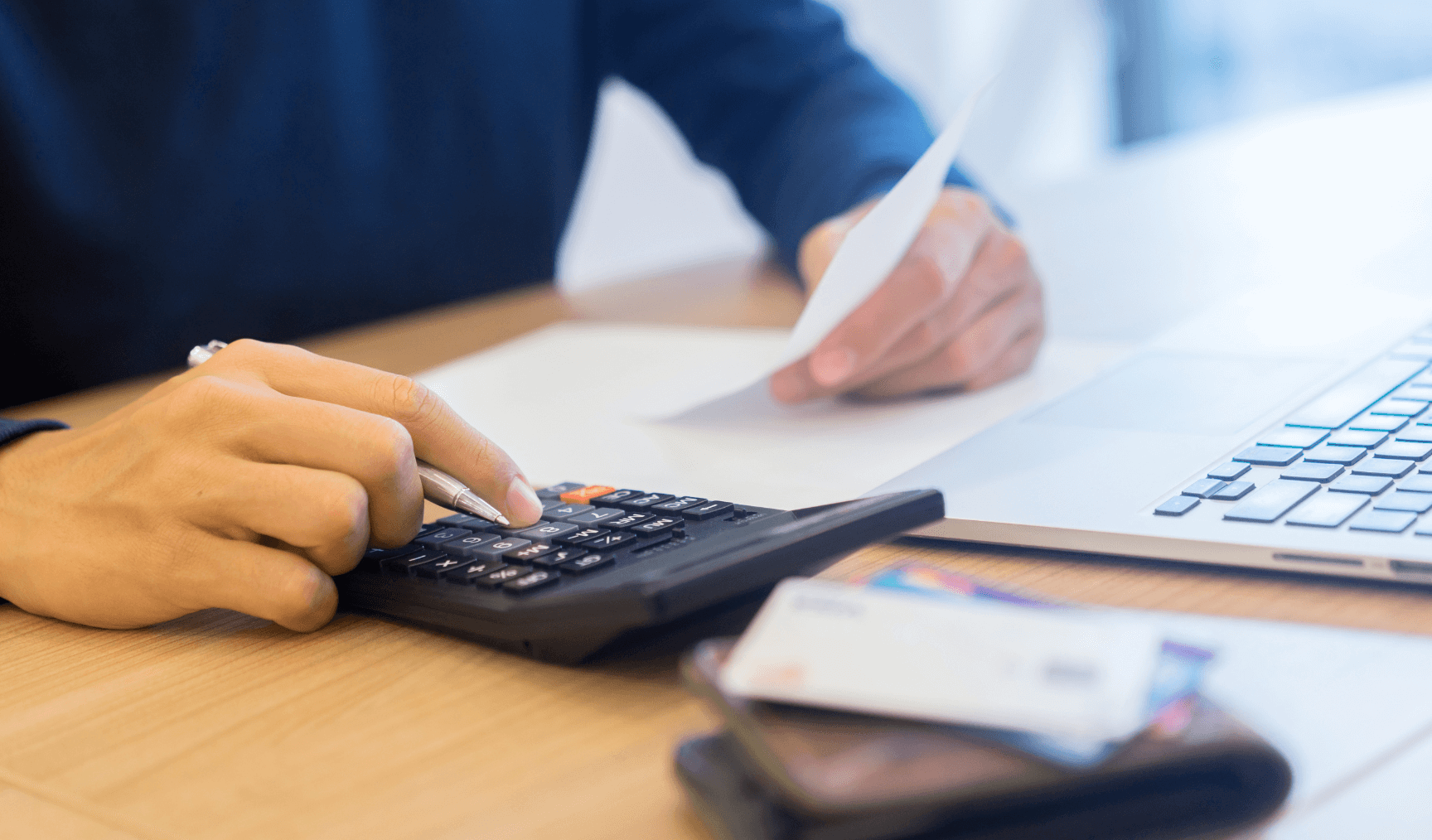 A person using a calculator and holding a piece of paper, working at a desk with a laptop in the background