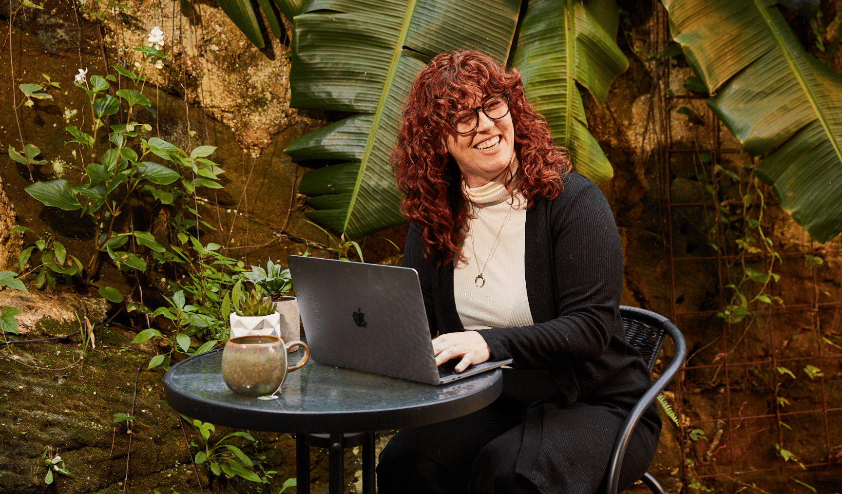 A woman with curly red hair and glasses, smiling while working on a laptop at an outdoor table surrounded by greenery