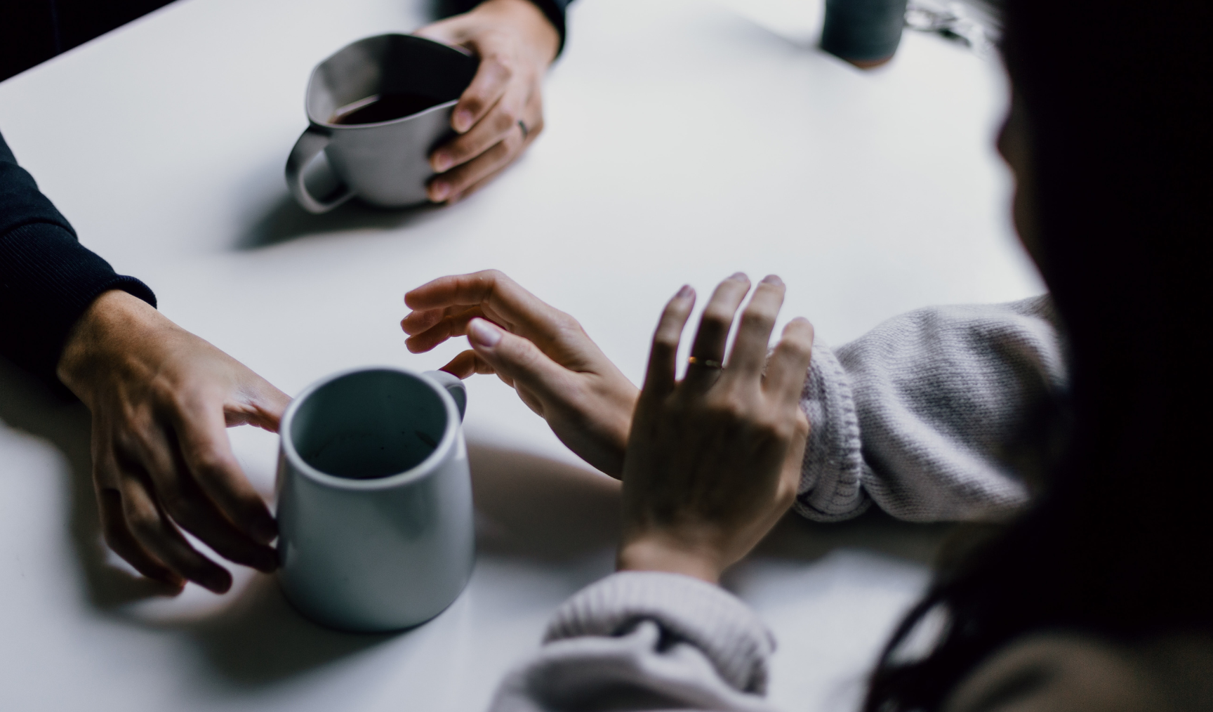 Two people sit at a table, only their hands visible. One is passing a mug of tea to the other