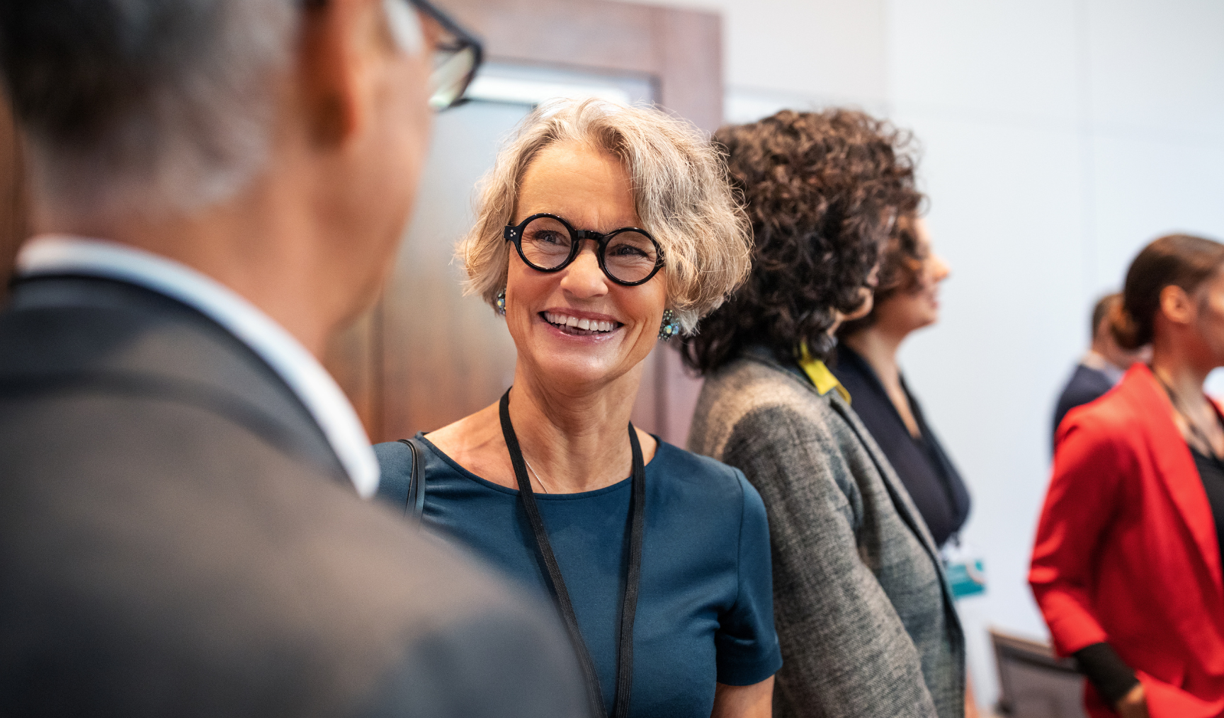 Smiling woman with gray curly hair and glasses in a business setting, engaging in conversation with colleagues