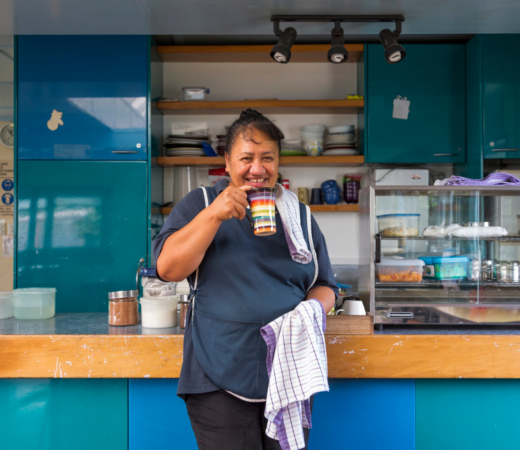 Smiling woman standing behind a counter in a kitchen, holding a mug and a dish towel, with shelves and kitchen equipment in the background