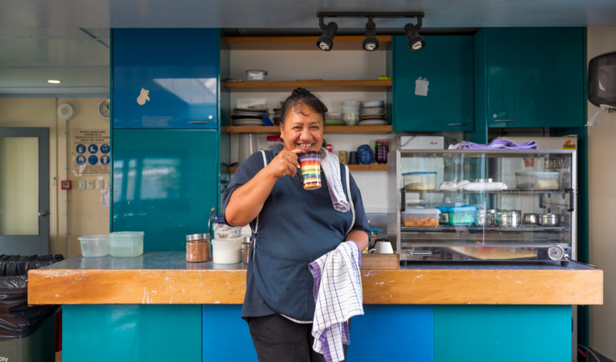 Smiling woman standing behind a counter in a kitchen, holding a mug and a dish towel, with shelves and kitchen equipment in the background