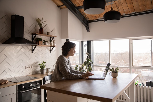 A woman, sitting at a wooden table in a modern kitchen, is working at a laptop. The kitchen has white subway tile backsplash, open shelves, and potted plants. There is a view of autumn trees from the large windows.