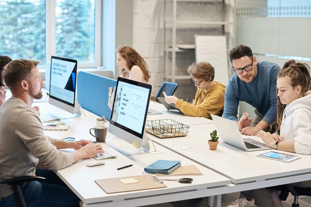 The image shows a modern, open-plan office space with several people working at a large white desk. A man and woman are seated on the left, focused on their computers, while another man assists the woman with her laptop. In the background, two women are working, with one partially obscured, and the desk is cluttered with office supplies, a collaborative and casual work environment.