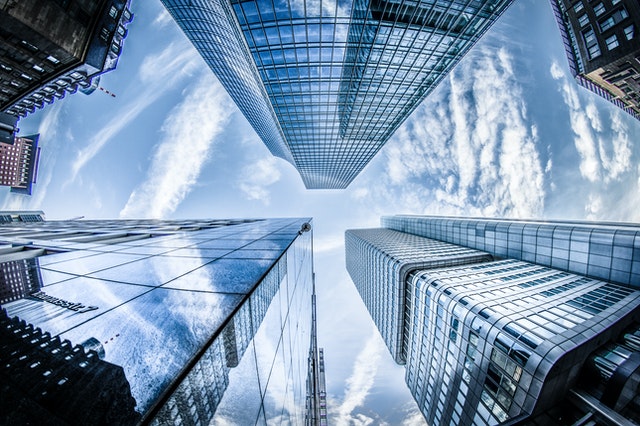 A photo taken from the ground looking up at the sky with skyscraper buildings towering above. There are also clouds reflected in the glass windows of the buildings.