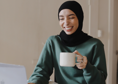 An image of a woman in hijab, smiling as she holds a mug while looking at her laptop