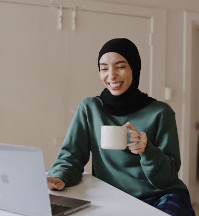 An image of a woman in hijab, smiling as she holds a mug while looking at her laptop