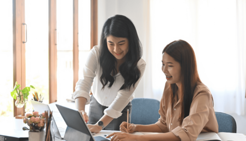 Two women working together at a desk, both smiling as they collaborate on a project. One is seated, taking notes, while the other stands, pointing to the laptop screen in a bright office environment