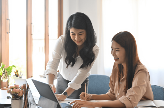 Two women working together at a desk, both smiling as they collaborate on a project. One is seated, taking notes, while the other stands, pointing to the laptop screen in a bright office environment