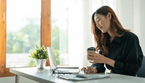 A woman sits at a desk in a bright office, holding a coffee cup while looking at a laptop