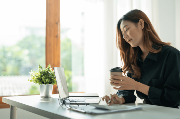 A woman sits at a desk in a bright office, holding a coffee cup while looking at a laptop