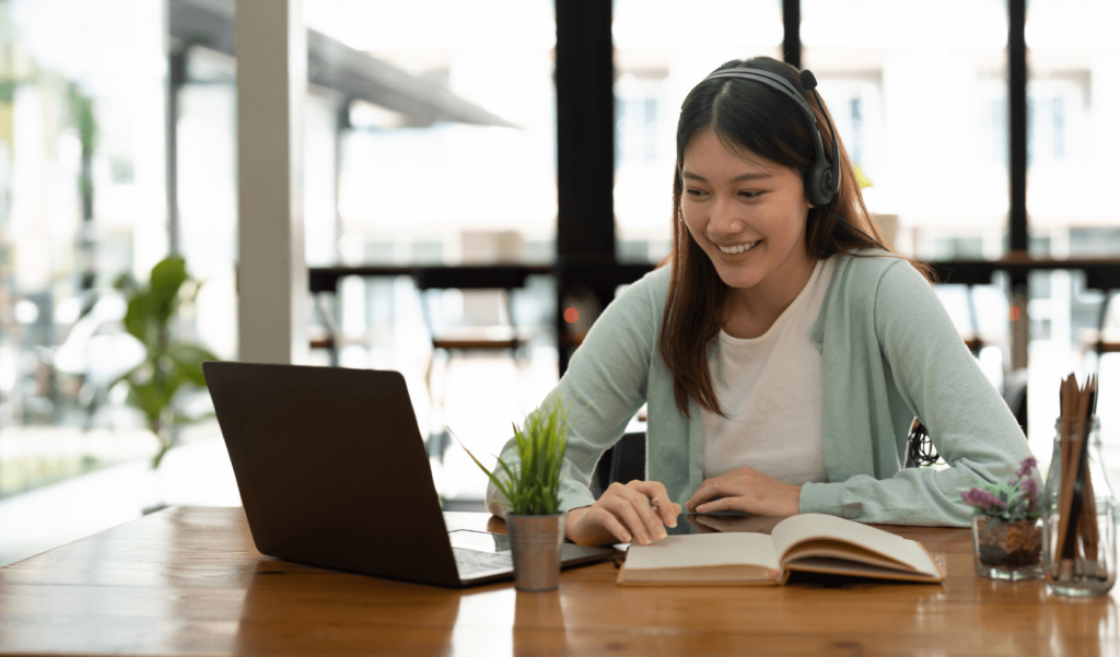 a woman wearing headphones and working at her laptop while smiling and reading a book