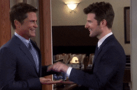 Two men in suits enthusiastically shaking hands in an office setting, smiling at each other with a look of excitement