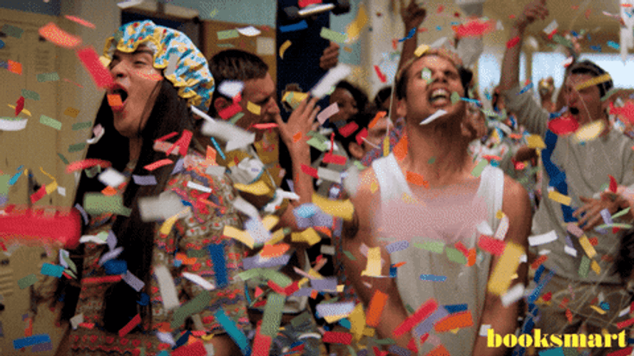 Group of people in a hallway celebrating with confetti, shouting joyfully, with the word 'booksmart' visible in the bottom right corner