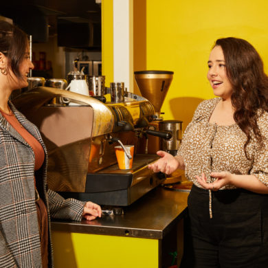 Two women having a conversation in a café setting near a coffee machine, with a bright yellow background.