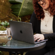 A woman sitting outdoors at a round glass table with a cup of coffee, working on a laptop while smiling. There are green plants in the background, creating a relaxed work environment.