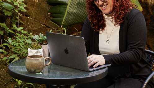 A woman sitting outdoors at a round glass table with a cup of coffee, working on a laptop while smiling. There are green plants in the background, creating a relaxed work environment.