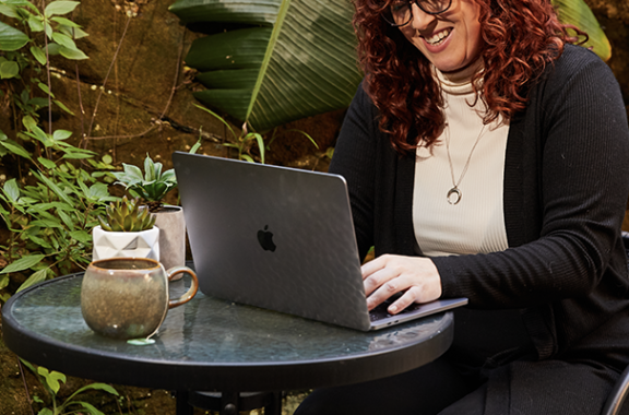 A woman sitting outdoors at a round glass table with a cup of coffee, working on a laptop while smiling. There are green plants in the background, creating a relaxed work environment.