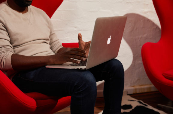 A person sitting on a red chair, working on a laptop. Only the lower part of the body and hands are visible, focusing on the laptop usage in a modern, comfortable workspace.
