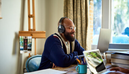 A man wearing headphones sits at a desk in a well-lit home office, engaged in a video call on his laptop, with books and a mug nearby.
