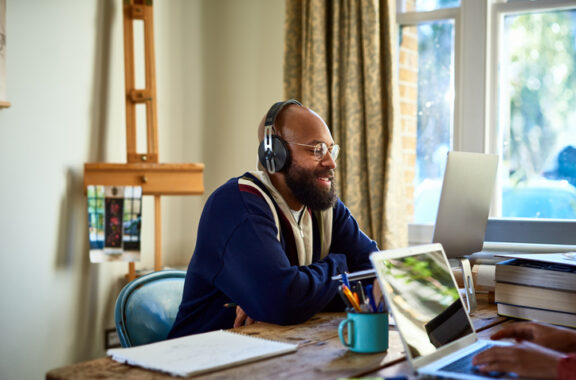 A man wearing headphones sits at a desk in a well-lit home office, engaged in a video call on his laptop, with books and a mug nearby.