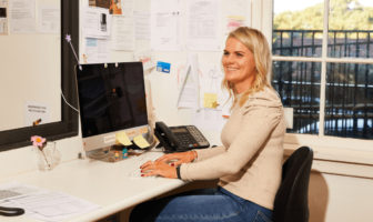 This image features a woman with blonde hair seated at a desk in an office setting, smiling as she faces a computer