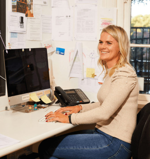 This image features a woman with blonde hair seated at a desk in an office setting, smiling as she faces a computer