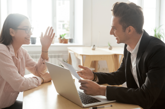 A professional man and woman engaging in a friendly discussion across a desk with a laptop, set in a bright office environment.