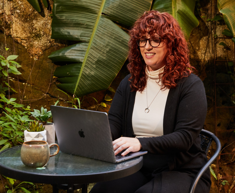 Person working on a laptop outdoors at a table, surrounded by lush green plants