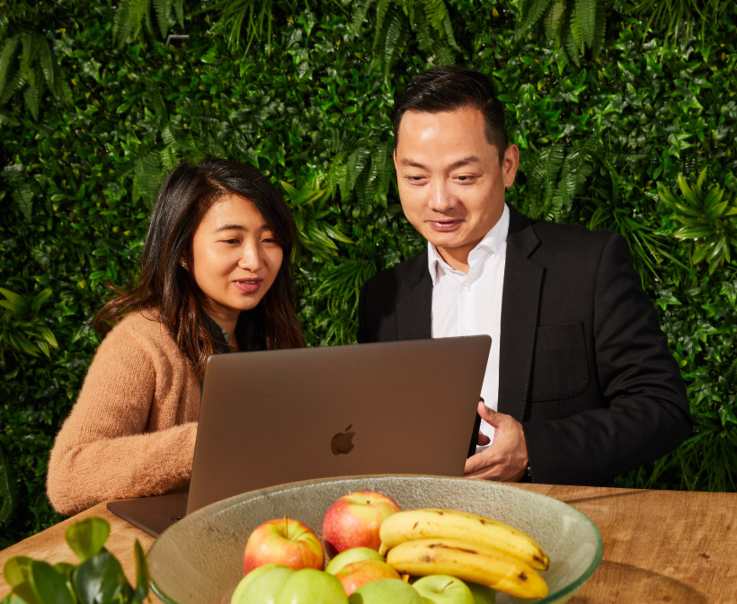 Two people looking at a laptop together, with a bowl of fresh fruit in the foreground and a lush green plant wall in the background
