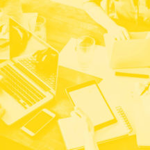 Group of people working collaboratively at a table with laptops, tablets, and notebooks, tinted in yellow