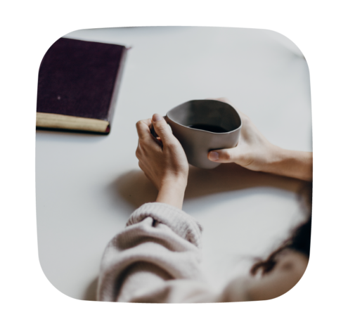A photo of a person holding a cup with both hands, sitting at a table with a closed book nearby