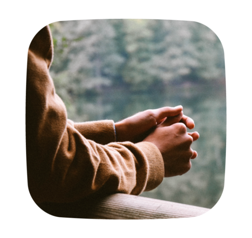 Close-up of a person's hands clasped together, resting on a wooden railing, with a blurred background of a calm lake and trees.