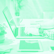 Person typing on a laptop at a desk with a tablet and a plant nearby, tinted in green