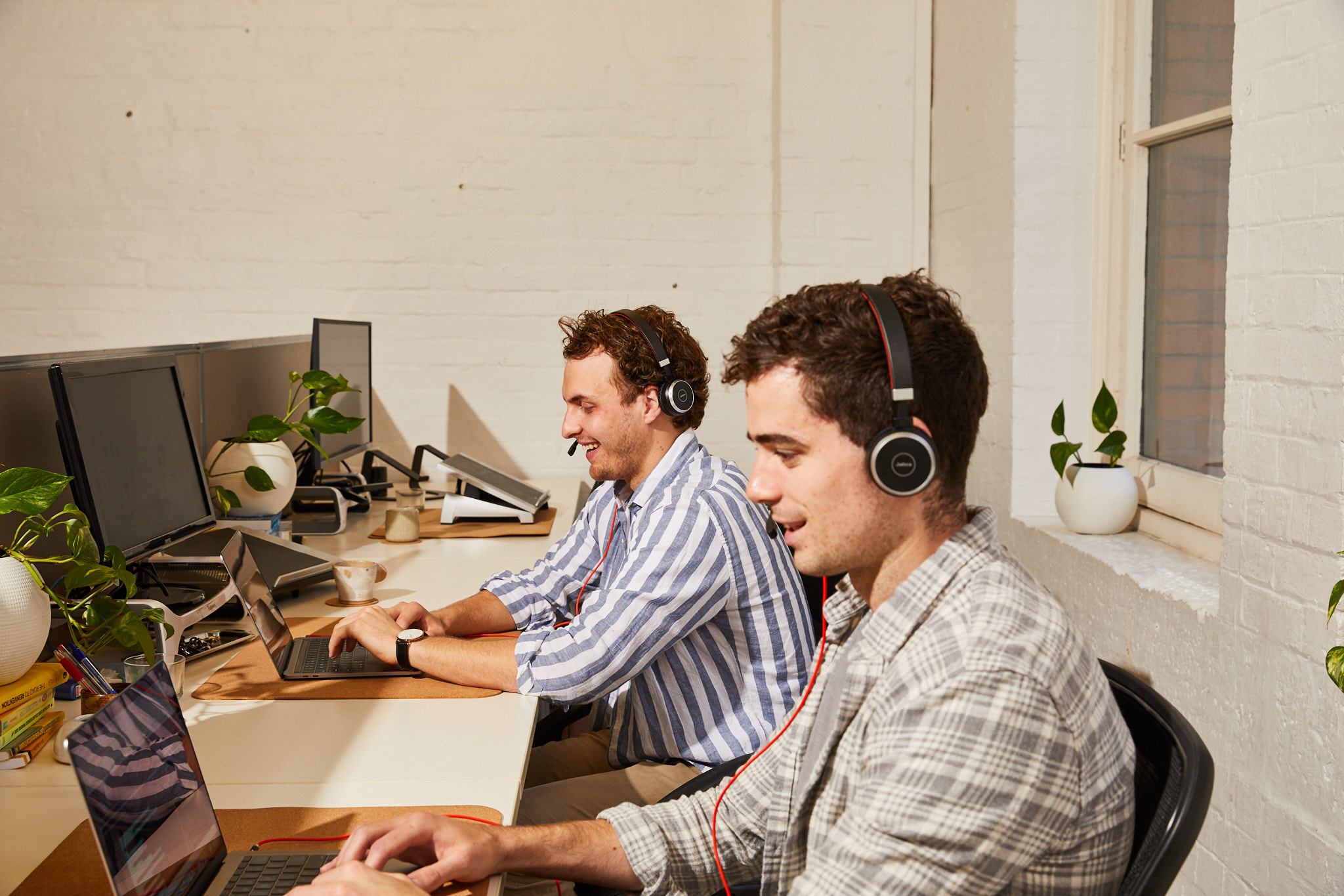 Two colleagues wearing headsets work at their desks, engaged in a conversation while using laptops