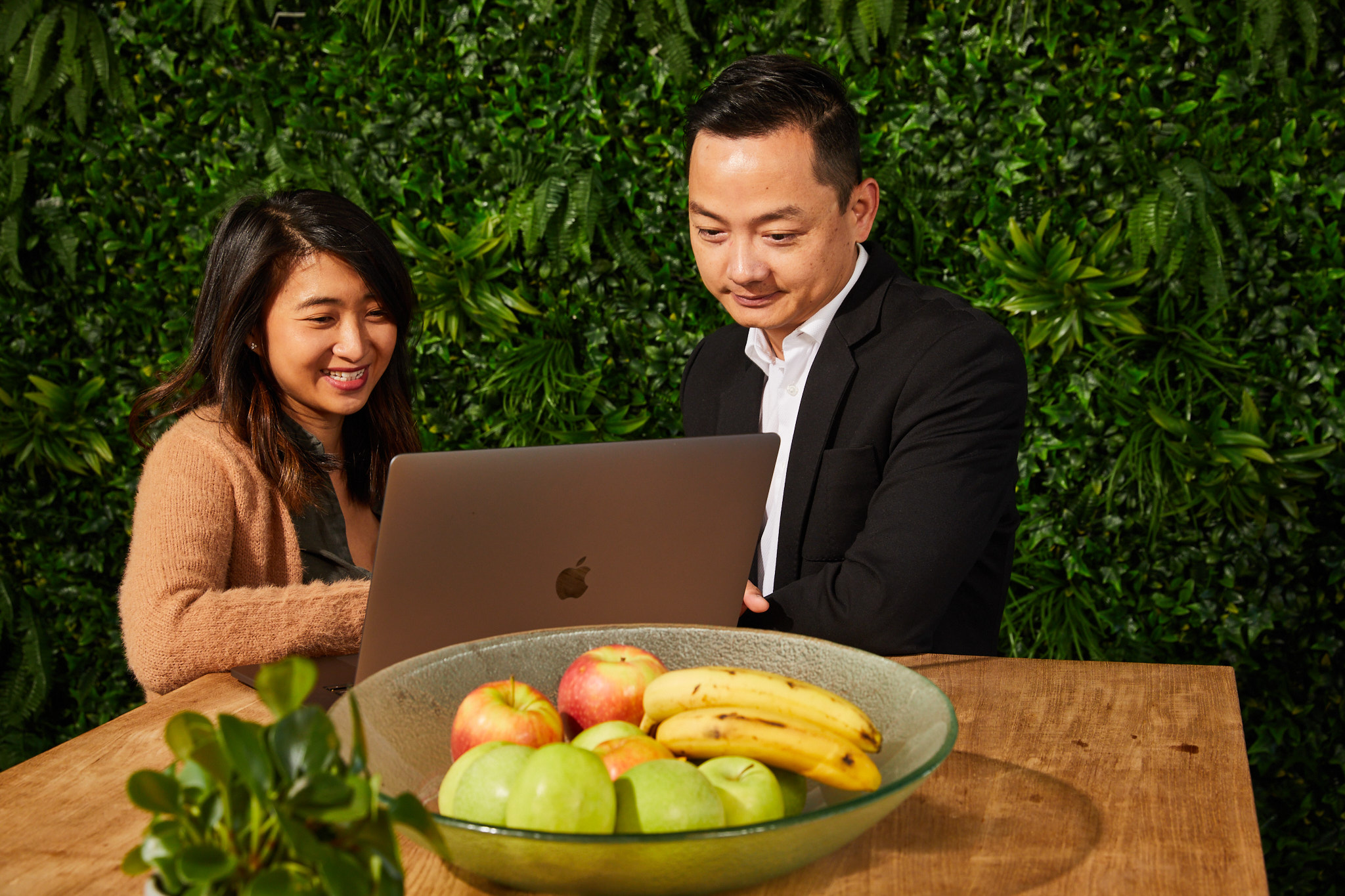 Two colleagues work on a laptop together, sitting at a table with fruit, against a vibrant green plant wall.