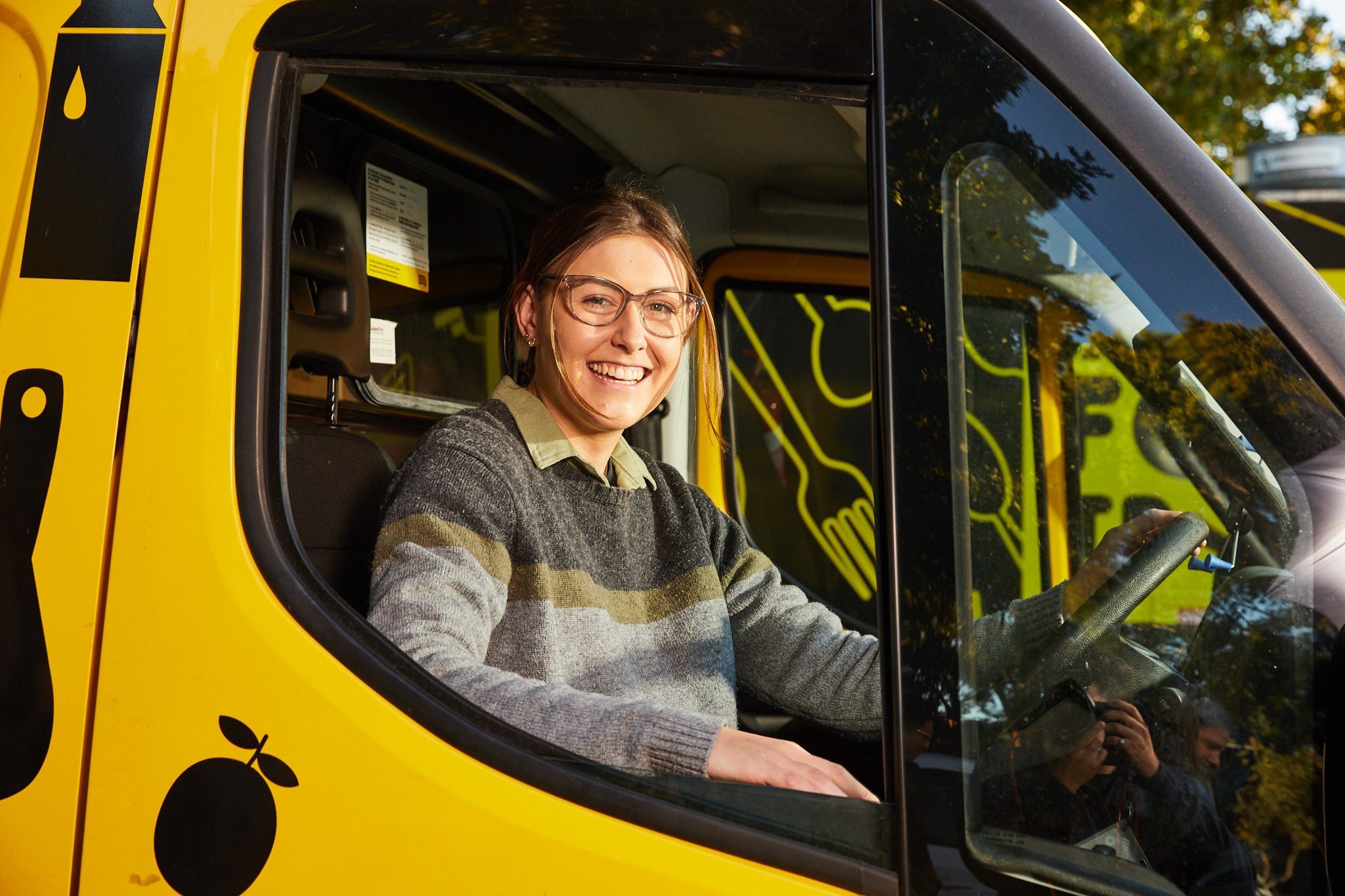 A woman sitting in the driver's seat of a yellow delivery truck, smiling at the camer