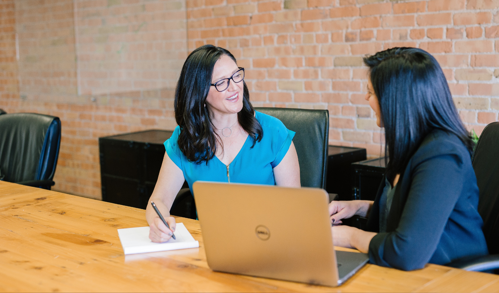 Two women sit at a wooden conference table, one taking notes, and both conversing, with a brick wall and black office chairs in the background. One has a laptop open.