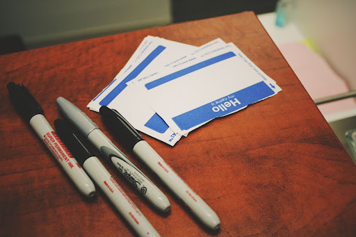 Stack of blank 'Hello, my name is' name tags on a wooden surface, accompanied by several permanent markers