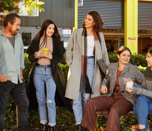 A group of six people casually standing and sitting outdoors, engaged in conversation and holding drinks, in a relaxed and friendly setting