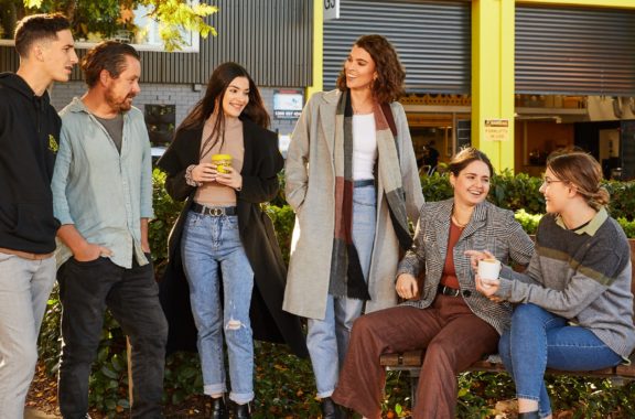A group of six people casually standing and sitting outdoors, engaged in conversation and holding drinks, in a relaxed and friendly setting