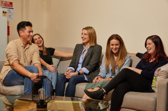 A group of five people sitting on a couch, laughing and enjoying a conversation in a relaxed, indoor setting
