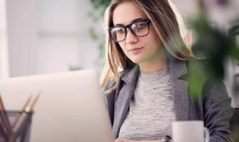 A woman wearing glasses and a blazer working on a laptop in a bright office environment