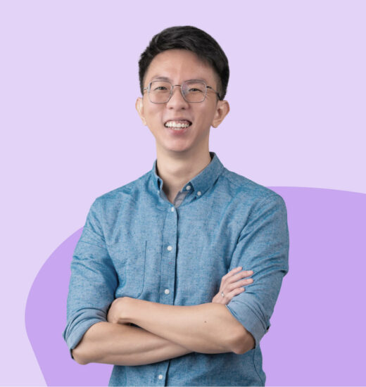 Smiling person with glasses, wearing a blue button-up shirt, standing with arms crossed against a light purple background