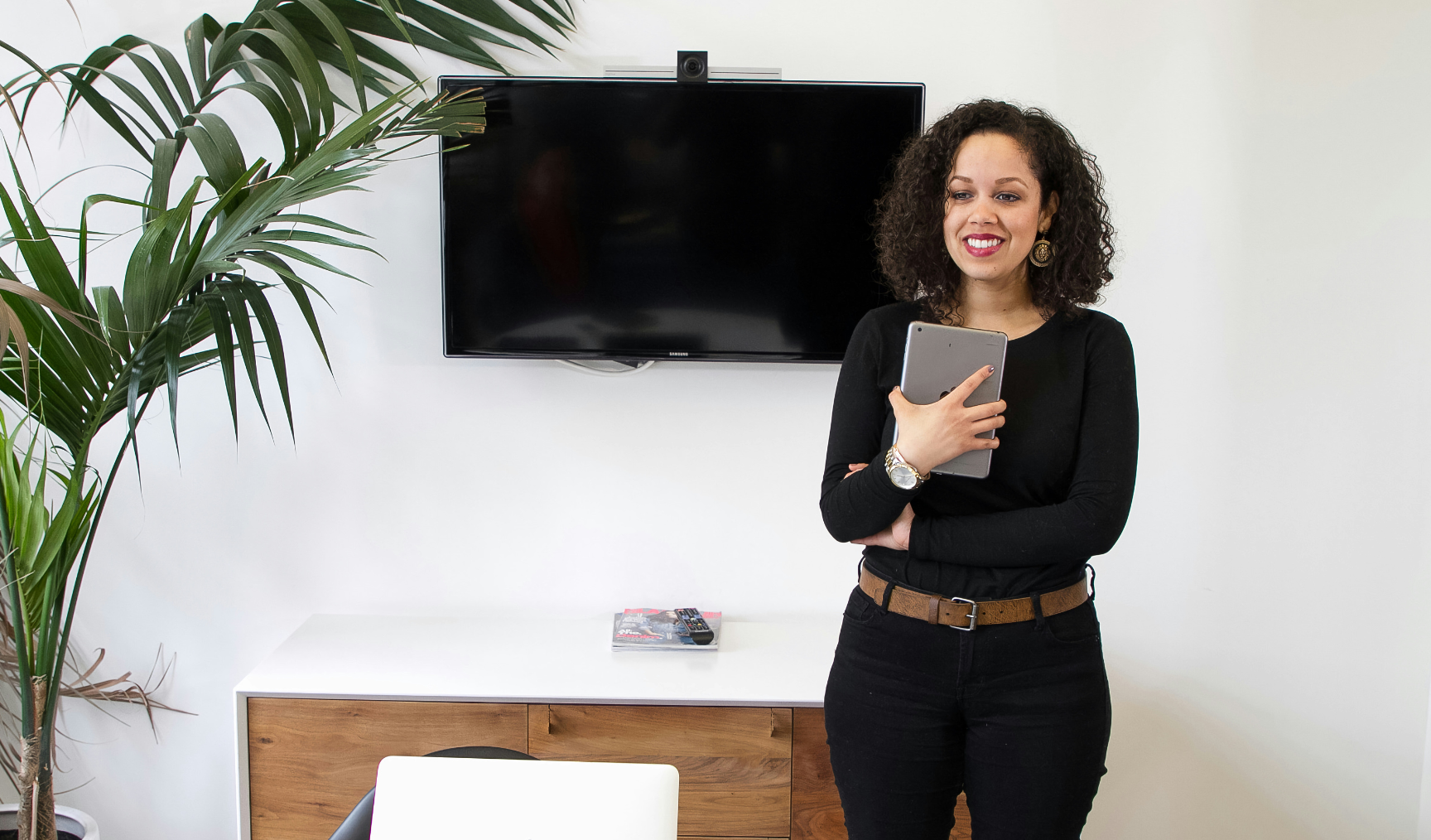 A woman stands at the head of a meeting room, tablet in hand, smiling