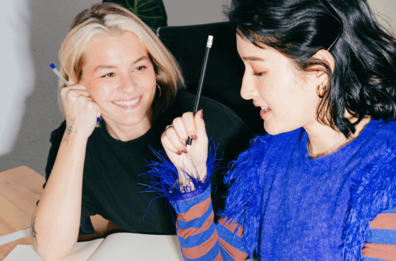 Two women sitting together, smiling and holding pens, engaged in conversation at a desk